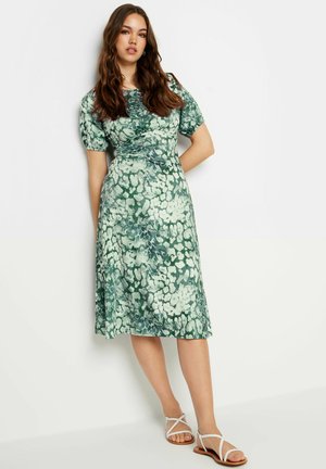 Vestido floral verde con mangas cortas abullonadas y detalles en la cintura, que presenta una falda fluida y una textura de tela suave. Las sandalias tienen tiras cruzadas.