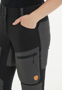 Zwarte en grijze cargobroek met een mix van gladde en gestructureerde stoffen, meerdere met rits afgesloten zakken en een klein bruin logo-embleem.