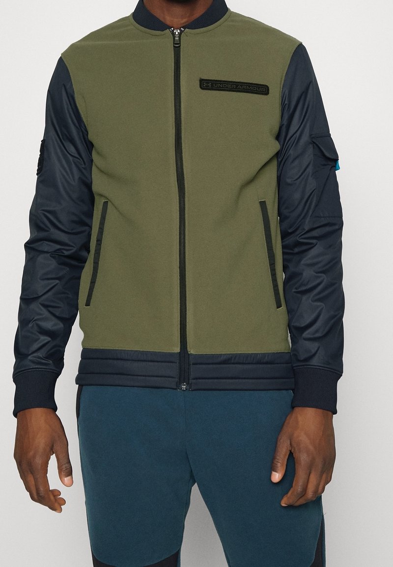 Béžově zelená bomber bunda s tmavě modrými rukávy, zipovým zapínáním, žebrovanými manžetami a lemem, s logem značky na hrudi a bočními kapsami.