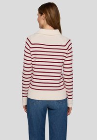 Cremefarbener Pullover mit burgunderfarbenen horizontalen Streifen, geripptem Strickmuster und einem Kragen. Wird mit blauen Jeans getragen und zeigt ein lässiges Design.