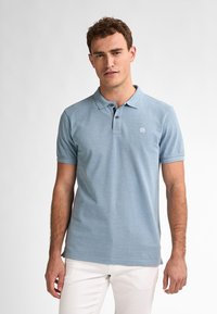 Světle modré polo tričko s klasickým límcem, krátkými rukávy a logem na levém prsu; v setu s bílými kalhotami. Hladká textura.