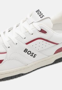 Bela usnjena športna obutev z rdečimi dodatki, teksturirano stransko ploščo, perforiranim dizajnom in črnim gumijastim podplatom. Na straneh ima črn logotip 'BOSS'.