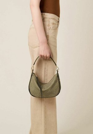 Donna con pantaloni beige e top marrone che tiene una piccola borsa verde oliva texturizzata con un manico corto al suo fianco.