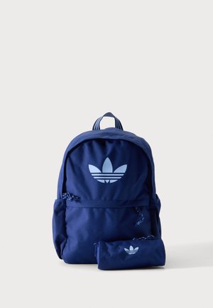 Sac à dos Adidas bleu avec poche avant zippée et trousse assortie, tous deux arborant un grand logo trèfle blanc, sur un fond clair uni.