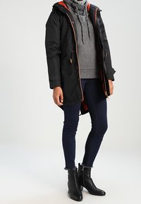 Czarna wodoodporna parka z pomarańczową podszewką, z kapturem, z zapięciem na zamek i kieszeniami bocznymi, noszona na szarym swetrze i ciemnych dżinsach.