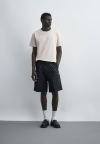 Homme debout portant une chemise beige à manches courtes, un short noir au genou avec une chaîne, des chaussettes blanches et des chaussures noires sur un fond gris uni.
