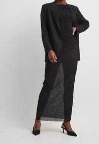 Schwarzer Blazer über einem strukturierten schwarzen Oberteil, kombiniert mit einem durchsichtigen schwarzen Rock. Das Ensemble zeichnet sich durch eine figurbetonte Silhouette und elegante Pumps aus.