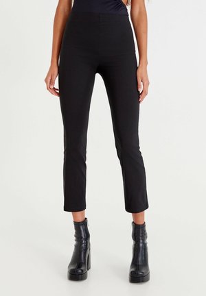 Femme portant un pantalon noir ajusté et court ainsi que des bottines noires à talons hauts, debout avec les bras détendus le long du corps.