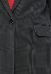 Blazer à carreaux noirs en tissu texturé, avec une fermeture à un bouton noir unique et une poche poitrine gauche. La doublure rouge est partiellement visible.