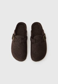 Brown felt clogs with a rounded shape, open back, and a side strap featuring a metal buckle. Soft texture and minimalist design.