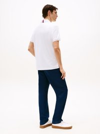 Man in een witte polo met rode en blauwe kraagstrepen, marineblauwe broek en witte sneakers met gumzolen, staande in profiel op een witte achtergrond.