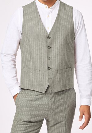 Homme portant un gilet gris clair à fines rayures et un pantalon assorti avec une chemise blanche boutonnée, mains partiellement dans les poches.