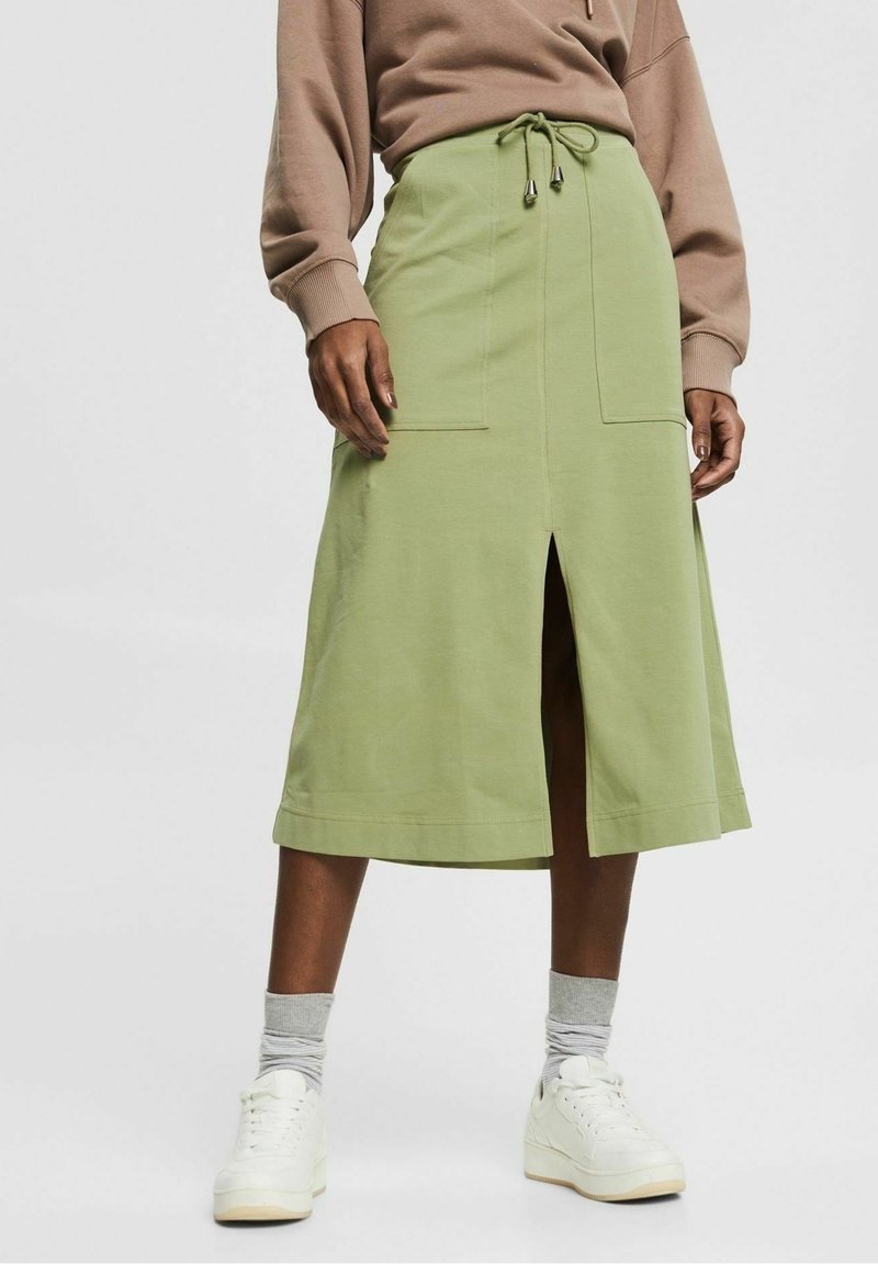 Femme portant une jupe midi vert clair avec une fente à l'avant, une taille à cordon, des poches plaquées, un sweat-shirt beige, des chaussettes grises et des baskets blanches.