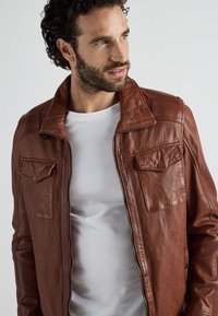 Brun läderjacka med en strukturerad design, två framfickor och dragkedjestängning. Bärs över en vit t-shirt med krage.