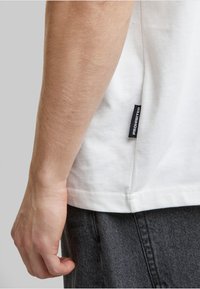 Weißes Baumwoll-T-Shirt mit geradem Schnitt. An der Seitennaht befindet sich ein kleines schwarzes Etikett mit der Aufschrift "VERBOTEN". Graue Denim-Hosen sind sichtbar.