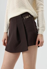 Bruine plissé short met een glanzende zilveren kettingaccent aan de taille, gedragen met een crèmekleurige gebreide trui met een ruitpatroon.