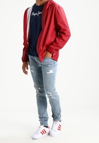 Röd lättviktig jacka över en marinblå T-shirt, slitna ljusblå jeans och vita sneakers med röda ränder. Fokus på avslappnad streetwear-stil.