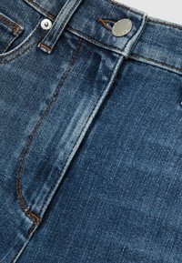 Jeans in denim blu scuro con un primo piano della vita con un bottone in metallo, cuciture a vista e sottili variazioni di texture.
