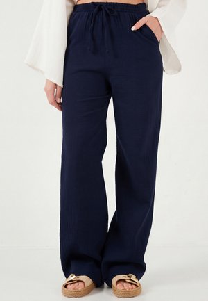 Granatowe szerokie spodnie z troczkiem w talii noszone z beżowymi sandałami na klamrę, zestawione z białą bluzką z długim rękawem, ręce spoczywają w kieszeniach.