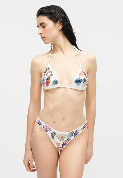 Femme aux cheveux mouillés portant un bikini blanc orné de motifs brodés colorés de coquillages et de coraux, sur un fond uni.