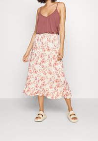 Femme portant un haut sans manches mauve, une jupe midi blanche avec un imprimé floral rose et un ourlet volanté, et des sandales beiges, se tenant contre un fond blanc.