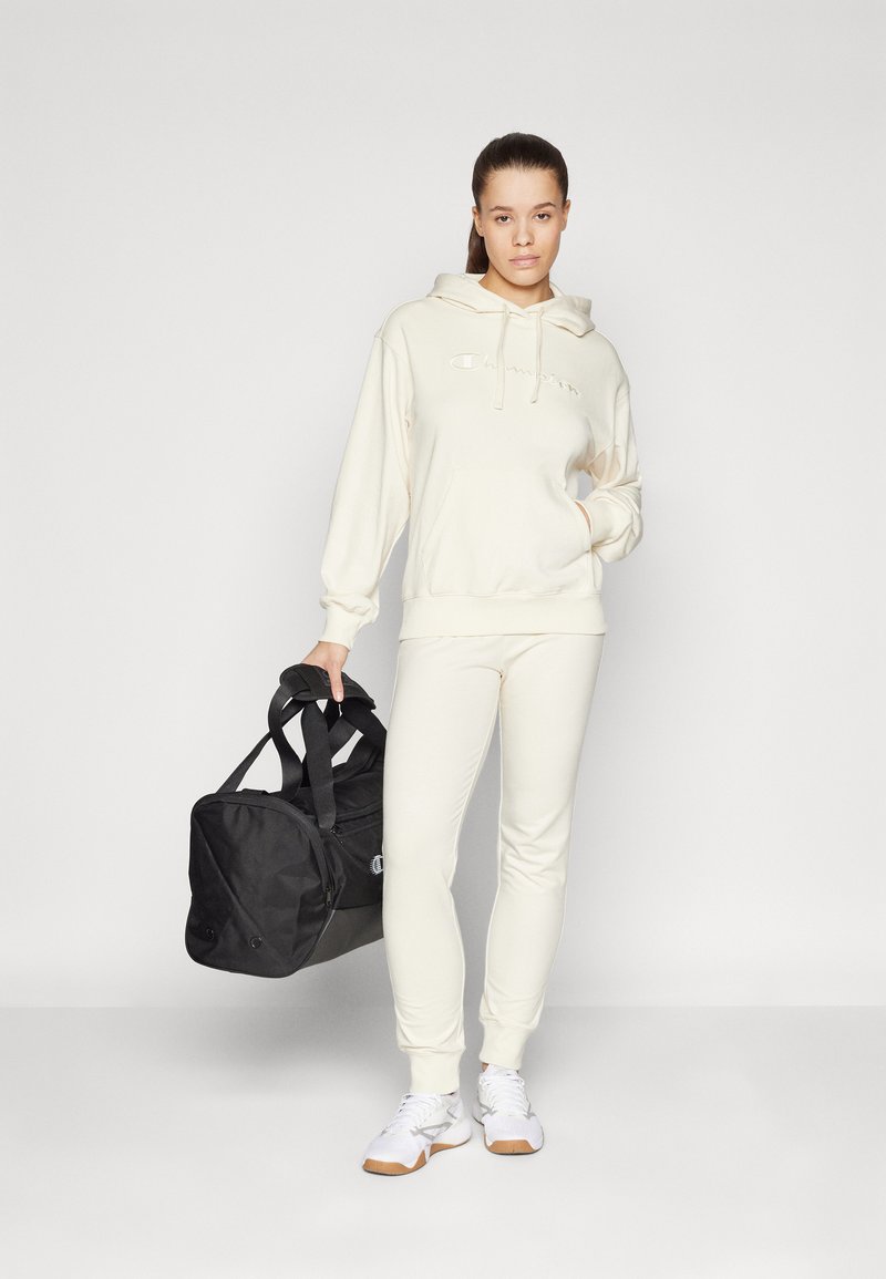 Sudadera crema con bolsillo frontal y logo bordado, combinada con pantalones a juego y zapatillas deportivas blancas, sosteniendo un bolso de deporte negro.
