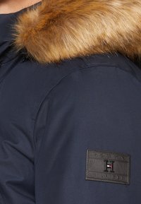 Námorně modrá zimní bunda Tommy Hilfiger s hnědým kožíškem na límci, s viditelným ramenem a logem značky na rukávu.