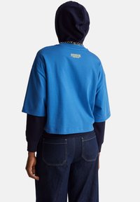 Modrá krátká mikina s námořnicky modrou kapucí, s logem "Stranger Things" ve žluté barvě na zádech, kombinovaná s tmavě modrými džínami.