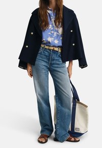 Cappotto doppiopetto navy sopra una camicia floreale azzurra chiara con motivi bianchi, abbinato a jeans a gamba larga azzurra chiara e sandali marroni. Borsa a tracolla in tela.