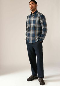Camisa de cuadros en tonos azul marino con líneas blancas y marrones, de algodón, de manga larga. Combinada con pantalones oscuros y zapatos brillantes marrones.
