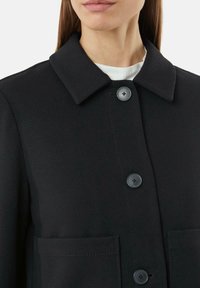 Czarny, teksturowany płaszcz z wywiniętym kołnierzem, z dwiema przednimi kieszeniami i czarnymi zapięciami na guziki. Prosty, klasyczny design.