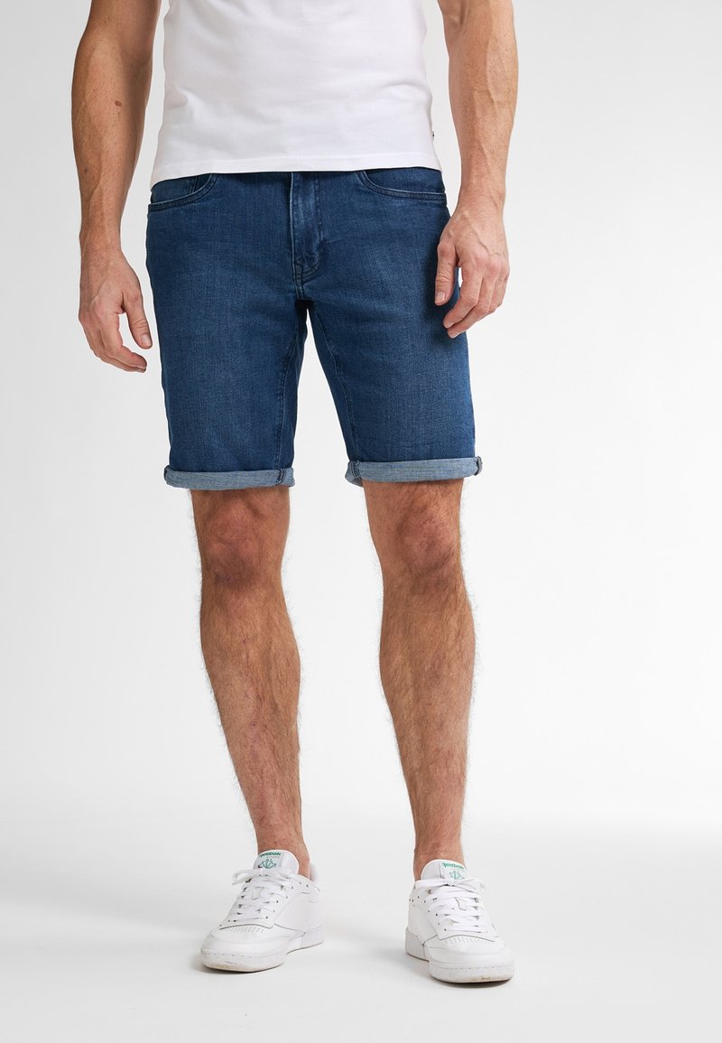 Shorts in denim blu con orli arrotolati, dotati di tasche anteriori e design aderente, indossati con sneakers bianche e una t-shirt bianca semplice.