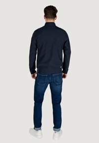 Giacca blu navy con colletto alto, slim fit e polsini a coste, abbinata a jeans blu scuro e sneaker bianche.