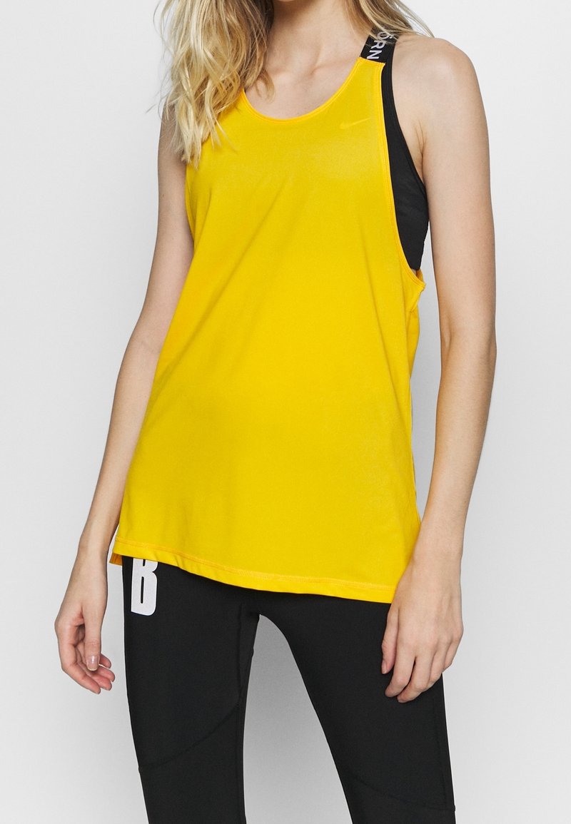 Żółta sportowa koszulka na ramiączkach o gładkiej fakturze, zaokrąglonym dekolcie oraz kontrastujących czarnych ramiączkach z logo. W połączeniu z czarnymi legginsami.