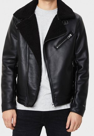 Chaqueta de cuero negra con grueso cuello de borreguito negro, cremallera asimétrica, bolsillo en el pecho con cremallera, llevada sobre una camiseta gris claro y pantalones negros.