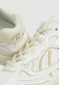 Zapatillas deportivas blancas que presentan una combinación de materiales de malla y cuero, con acentos en crema y un diseño curvado, atadas con cordones planos y texturizados.