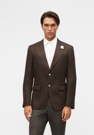 Hombre vestido con una chaqueta marrón oscura, camisa blanca y pantalones grises, de pie frente a un fondo liso de color gris claro.