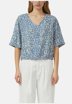 Femme portant un chemisier bleu et blanc à pois, col en V, manches courtes, légèrement rentré dans un pantalon blanc plissé taille haute.