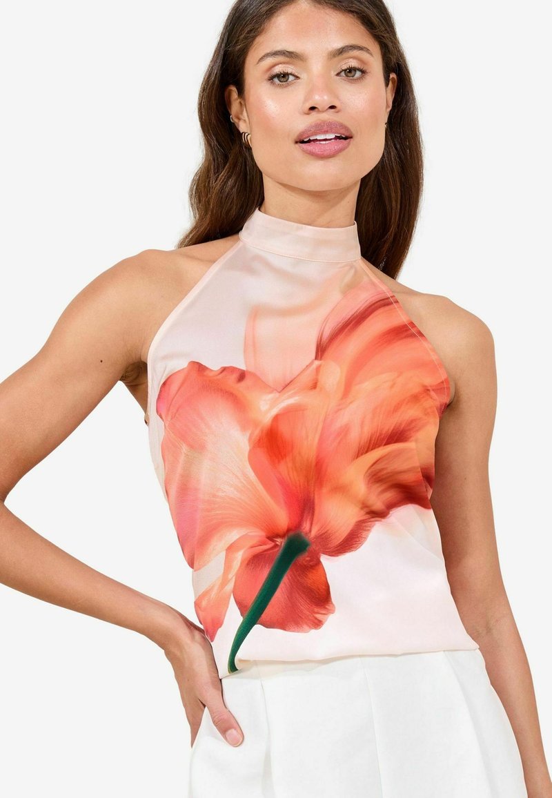Květinový halter top s potiskem růžových a oranžových květin na jemné tkanině, s vysokým výstřihem a hladkým povrchem. Na sobě má bílé kalhoty.