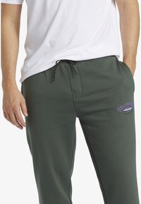 Donkergroene sweatpants met elastische tailleband en zwarte trekkoord, met een paarse logo aan de linkerzijde en zijzakken.