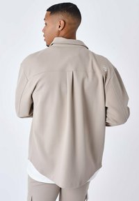 Beige langærmet skjorte med krave, der har et struktureret stof, en læg bagpå og et diskret logo på skulderen.