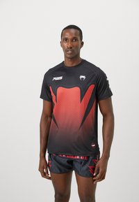 Schwarzes sportliches Hemd mit einem roten Farbverlauf-Design, das ein strukturiertes Muster und ein Logo enthält. Kombiniert mit schwarzen Shorts mit roten Akzenten.