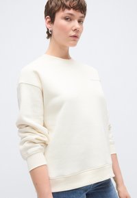 Krēmkrāsas džemperis ar apaļu kakla izgriezumu, ribotiem aprociem un apakšmalu. Mīksta tekstūra ar smalku logotipa detaļu krūšu daļā.