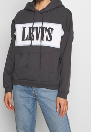 Person iført en sort hættetrøje med hvidt panel og stor sort "LEVI'S" tekst, kombineret med lyseblå jeans, stående foran en ensfarvet baggrund.