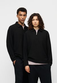 Μαύρο πουλόβερ μακρυμάνικο με γιακά και κλείσιμο με μισή φερμουάρ. Μαλακή υφή, άνετη εφαρμογή, συνδυασμένο με σκούρο παντελόνι. Μινιμαλιστικός σχεδιασμός.