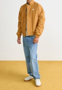 Senapsgul lätt jacka med hög krage, tryckknappar och elastiska ärmmuddar, bärs över en ljusblå skjorta och medelblå jeans.