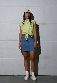 Camisa sem mangas verde-neon com atilho à frente; saia de ganga azul com bainha desfiada; sapatilhas altas brancas com plataforma; boné bege com logótipo.