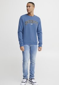 Sudadera azul con el texto "UNIVERSITY", combinada con vaqueros de un azul claro y zapatillas blancas con rayas negras.