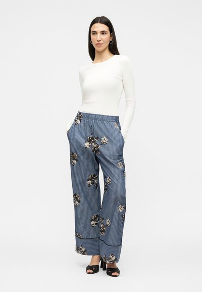 Femme debout, les mains dans les poches, portant un haut blanc à manches longues, un pantalon large à rayures bleues avec imprimé floral et des talons ouverts noirs.
