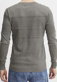 Persona che indossa un maglione grigio a maniche lunghe con texture e jeans blu, mostrata di spalle con le braccia rilassate lungo i fianchi.